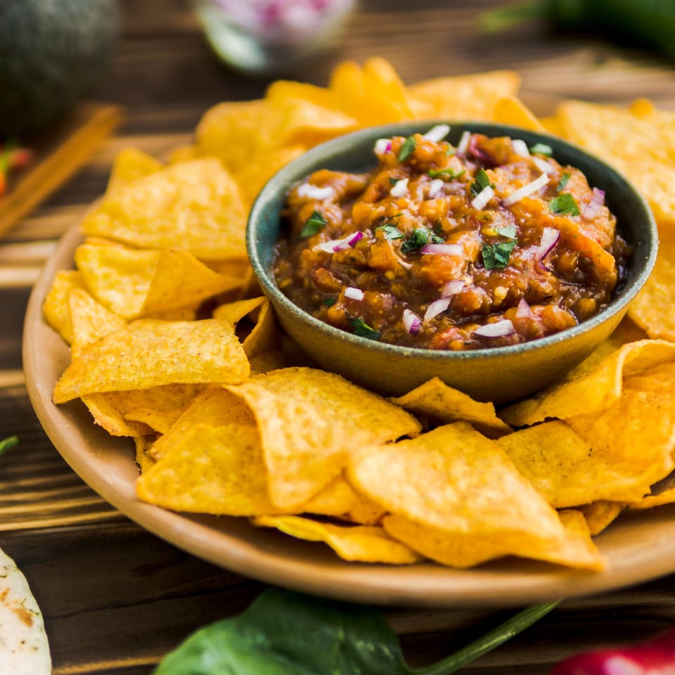 A bowl of chunky salsa with red onion and cilantro, surrounded by tortilla chips on a wooden plate.